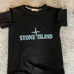 Svart Stone Island t-shirt med tryck - Svart t-shirt från Stone Island med stort ljusblått logotryck på bröstet. Klassisk rund hals och korta ärmar. Tillverkad i mjuk bomull för en skön känsla och clean look.