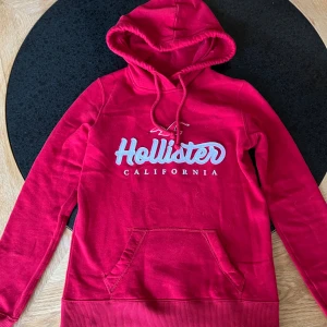 Röd hoodie från Hollister California - Snygg röd hoodie från Hollister California med vit logotyp på bröstet. Tröjan har huva med snörning, magficka och ribbade muddar vid ärmslut och nederkant. Perfekt för dig som gillar streetwear och vill ha en chill look.