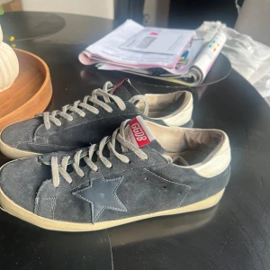 Golden goose mörkblå/blå  st44 - Golden Goose sneakers i mörkblå mocka med vit läderdetalj på hälen och en ikonisk stjärna på sidan. Skorna har snörning, rund tå och beige platt sula. GGDB-logga på tungan och sidan. Perfekt för dig som gillar en cool och avslappnad stil. Skorna är i storlek 45 men passar 44. I tån och hälen har skon släppt (ses på bild) detta kan fixas enkelt med lite skolim. Därav priset. Hör av er vid funderingar 