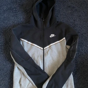 Nike hoodie svart och grå XS - Nike hoodie i svart och grått med vit logga på bröstet. Tröjan har hel dragkedja, huva och en snygg vit linje över bröstet. Långärmad modell med ficka på ärmen och normal passform. Perfekt för chill eller träning.