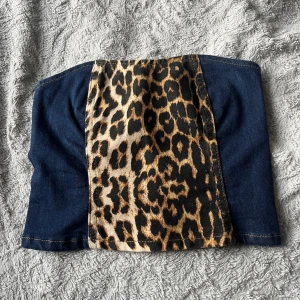 Tubtopp i denim med leopardmönster - Snygg tubtopp i mörkblå denim med en mittpanel i leopardmönstrat tyg. Toppen är axelbandslös och har en cool kontrast mellan det blå jeanstyget och det djuriska mönstret framtill. Perfekt för dig som vill sticka ut med en edgy och trendig look.