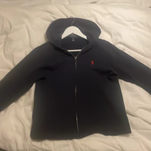 Svart hoodie från Polo Ralph Lauren - Marin blå hoodie från Polo Ralph Lauren med dragkedja och klassisk röd broderad logga på bröstet. Tröjan har huva och är långärmad, perfekt för en chill och stilren look. Tillverkad i mjukt material som känns skönt mot huden.