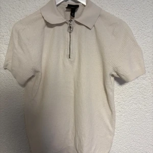 Vit kortärmad T-shirt med dragkedja H&M - Säljer en vit kortärmad T-shirt från H&M med krage och snygg silverfärgad dragkedja framtill. Toppen har en diskret struktur i tyget och är i slim fit-modell. Perfekt för dig som gillar stilrena och enkla plagg med en twist.