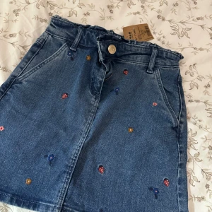 Blå jeanskjol med broderade blommor - Söt blå jeanskjol från Name It med broderade små blommor i olika färger. Kjolen har klassisk femficksdesign, bälteshällor och stängs med knapp och dragkedja framtill. Perfekt för dig som gillar denim med en twist!