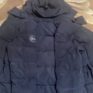 Canada Goose black label - Säljer en svart dunjacka från Canada Goose med huva och diskret logga på bröstet. Jackan har en quiltad design, knappar framtill och är perfekt för kalla dagar. Klassisk och stilren modell som håller dig varm hela vintern.