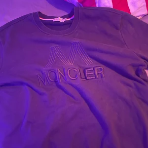 Svart Moncler sweatshirt 3XL - Svart sweatshirt från Moncler i storlek 3XL med stort broderat Moncler-logo på bröstet och klassisk rund hals. Tröjan har långa ärmar och en liten Moncler-patch på ena ärmen. Tillverkad i mjuk bomull för en skön och avslappnad stil.
