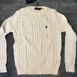 Vit kabelstickad tröja Polo Ralph Lauren - Vit kabelstickad tröja från Polo Ralph Lauren i storlek M. Klassisk rund halsringning och ribbade muddar. Tröjan har det ikoniska Polo-logot broderat på bröstet i mörkblått. Perfekt för dig som gillar stilren och tidlös design.