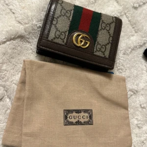 Gucci plånbok med GG-logga och ränder - Snygg plånbok från Gucci i brunt skinn med klassiskt GG-monogram och gröna/röda ränder. Guldigt GG-emblem framtill och flera kortfack inuti. Perfekt accessoar för dig som gillar lyxiga detaljer och ikonisk design.
