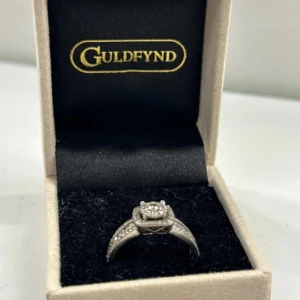 Diamant ring i vitt guld  - Snygg silverfärgad ring från Guldfynd med glittrande stenar i en fyrkantig design. Ringen har dubbla band med små stenar längs sidorna och en större sten i mitten, vilket ger en lyxig och modern look. Perfekt för dig som gillar bling och stil.