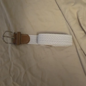 Vitt flätat bälte med skinn-detaljer 105 cm - Snyggt vitt bälte med flätad design och bruna skinn-detaljer vid spännet. Spännet är i silverfärgad metall och bältet har en stilren look som passar till många outfits. Perfekt accessoar för att lyfta din stil. 105 cm