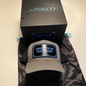 Grå keps John Hatter & Co - Snygg grå keps från John Hatter & Co med svart mesh på sidorna och bak. Framtill sitter en metallskylt med texten 'Gentleman by day, Beast by night' i blått. Perfekt statement piece med cool design och justerbar passform. En liten fläck på en sidan men annars fin syns inte bara om man tänker på det.