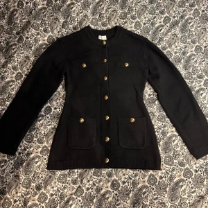 Svart kofta med guldknappar från H&M - Snygg svart kofta från H&M med guldiga knappar framtill och på fickorna. Modellen är långärmad och har två stora fickor framtill. Koftan är stickad och har en klassisk, lite längre passform som ger en stilren look. Koftan är i storlek XS men passar även de som bär S om de vill ha en figurnära look. (Jag på bilden bär vanligtvis S). Använd vid ett tillfälle. 