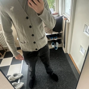 Grå stickad kofta Abercrombie & Fitch - Snygg grå stickad kofta från Abercrombie & Fitch med v-ringning och svarta knappar framtill. Västen har två fickor och en vit broderad älg på bröstet. Perfekt för lager-på-lager och chill stil. Ribbad kant vid ärmslut och nederkant.