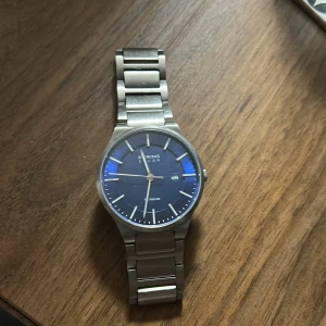 Bering Solar klocka i titan - Stilren Bering Solar klocka med blå urtavla och datumvisning. Boett och länk i matt titan ger en modern känsla. Klockan har silverfärgade index och visare, samt solcellsdriven urverk. Perfekt för dig som gillar minimalistisk design.