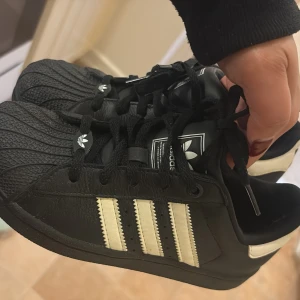 Svarta Adidas Superstar sneakers - Klassiska svarta Adidas Superstar sneakers med vita ränder och snörning. Skorna har den ikoniska gummitå-designen och är tillverkade i skinn med detaljer i syntet. Perfekta för en streetstyle-look.