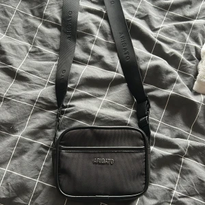 Svart axelväska från Arigato - Snygg svart axelväska från Arigato med justerbar bred rem där loggan är broderad. Väskan är väldigt stilren