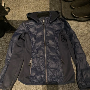 Mörkblå från JH Collection - Mörkblå jacka med huva från JH Collection. Jackan har dragkedja framtill, två fickor med dragkedja och är tillverkad i en mix av polyester och fleece. Snygg quiltad look på framsidan och ärmarna. Hårstråna tas bort innan den skickas och smutsen på ärmarna går säkert bort i tvätten! 