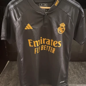 Real Madrid svart bortatröja Adidas S - Svart Real Madrid bortatröja från Adidas i slim fit. Tröjan har korta ärmar, guldfärgade detaljer och klubbmärke, samt 'Emirates Fly Better' tryckt på bröstet. Tillverkad i lätt och ventilerande material, perfekt för fotboll eller att bära som supporter.