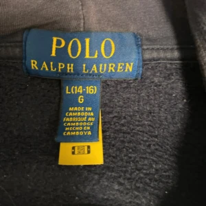 Mörkblå hoodie från Polo Ralph Lauren - Mörkblå hoodie från Polo Ralph Lauren med dragkedja och huva. Klassisk röd broderad logga på bröstet och kängurufickor framtill. Perfekt för en avslappnad stil och enkel att matcha med jeans eller joggers.