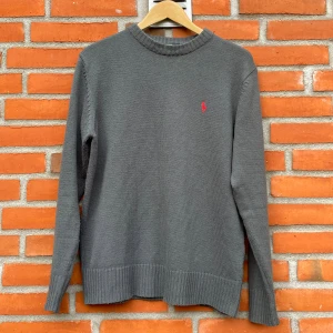 Grå stickad tröja från Ralph Lauren - Klassisk grå stickad tröja från Polo Ralph Lauren med den ikoniska röda loggan på bröstet. Tröjan har en tidlös design med rund hals och ribbade muddar som ger en perfekt passform. Perfekt för både vardag och mer uppklädda tillfällen – snygg att bära över en skjorta eller ensam för en avslappnad look. I mycket fint skick utan synliga defekter. Storlek: storlek M, axelbredd 43 och längd 67 cm  Färg: Mörkgrå med röd logga Passform: Normal Material: Antar att det är ull eller bomull, inte säker  