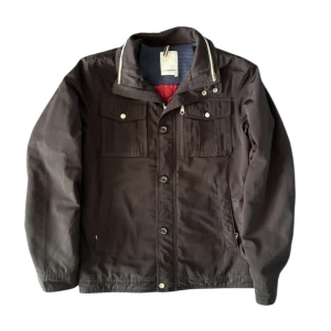 J.lindeberg field jacket svart quiltad jacka - Stilren svart fieldjacket från J.Lindeberg med quiltat rött foder. Jackan har flera fickor med knappar, dragkedja och knappstängning framtill samt detaljer vid kragen. Perfekt för höst och vår, med en snygg och funktionell look.