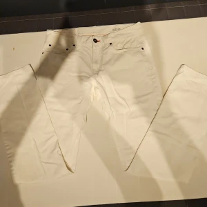 Vita straight fit jeans Hudson W31 L34 - Snygga vita jeans från Tommy Hilfigerl i straight fit-modell. Klassisk femficksdesign med svarta nitar och detaljer, samt läderpatch och liten logga bak. Tillverkade i bomull för en skön känsla och stilren look.