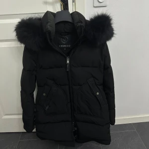 Svart pufferjacka från Cedrico med äkta päls - Snygg svart pufferjacka från Cedrico med stor huva och fluffig äkta päls. Jackan har dragkedja framtill, två stora fickor med knappar och detaljerade sömmar. Perfekt för kalla dagar när du vill hålla stilen och värmen. 