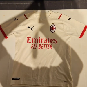 AC Milan kortärmad matchtröja Puma - AC Milan officiell matchtröja från Puma i ljus beige med röda detaljer och klubbmärke på bröstet. Tröjan har korta ärmar, rund hals och ventilerande meshpaneler. Materialet är lätt och andas, perfekt för fotboll. Emirates sponsortryck på framsidan. Den har lite skador på magen som man kan se på sista bilden och en fett fläck men annars är tröjan i bra skick.