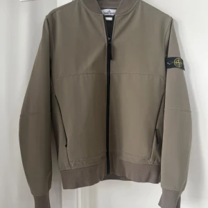  Stone Island jacka - Snygg beige/grön jacka från Stone Island med klassisk patch på ärmen. Jackan har ribbade muddar, dragkedja framtill och två fickor med dragkedja. Storlek XS/170