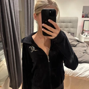 Svart Juicy Couture hoodie med strass - Säljer en svart hoodie från Juicy Couture i mjuk velour med strassdetaljer på bröstet och stor Juicy Couture-logga i strass på ryggen. Tröjan har huva, dragkedja och fickor framtill. Perfekt för dig som gillar bling och klassisk Y2K-stil.