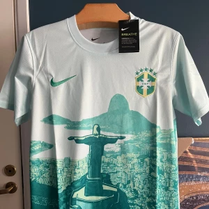Brasilien fotbollströja Nike S - Snygg Brasilien fotbollströja från Nike i ljusblått med grönt tryck av Kristusstatyn och Rio de Janeiro. Kortärmad modell med rund hals och CBF-emblem på bröstet. Tillverkad i lätt, ventilerande Dri-FIT material som håller dig sval under matchen.