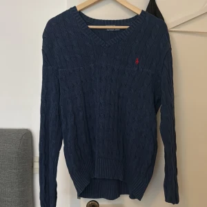 Marinblå kabelstickad tröja Polo Ralph Lauren - Kabelstickad marinblå tröja från Polo Ralph Lauren med röd broderad logga på bröstet. V-ringad modell med långa ärmar och ribbade muddar. Perfekt för lager-på-lager och klassisk preppy stil.