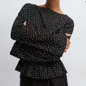 Svart prickig blus med volang - Säljer en svart blus med vita prickar och volangkant nedtill. Blusen har långärmade, lätt utsvängda ärmar och smockad midja som ger en snygg siluett. Materialet är mjukt och lätt, perfekt för dig som gillar trendiga detaljer och mönster.