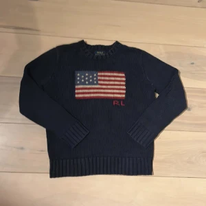 Ralph lauren tröja 🤴🏼 - Snygg mörkblå stickad tröja från Polo Ralph Lauren med amerikansk flagga broderad på bröstet och RL-detalj i rött. Tröjan har rund hals, långa ärmar och breda ribbade muddar vid ärmslut och nederkant. Perfekt för dig som gillar klassisk stil med en twist.