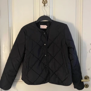 Svart quiltad jacka från Nelly - Stilren svart quiltad jacka från Nelly med rund halsringning och tryckknappar framtill. Jackan har lång ärm och ett rakt snitt, perfekt för lager-på-lager. Materialet är lätt och känns smidigt, vilket gör den enkel att bära under höst och vår. Jackan är sparsam använd och i ett jättebra skick! Hör av er för frågor osv.