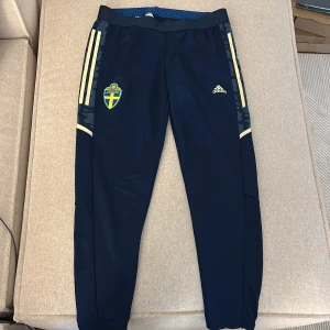 Adidas Sverige träningsbyxor marinblå - Marinblå träningsbyxor från Adidas med Sveriges landslagsemblem på ena benet och klassiska gula ränder längs sidorna. Byxorna har smal passform, elastisk midja och är tillverkade i ett syntetmaterial som andas. Perfekta för fotbollsträning eller chill.