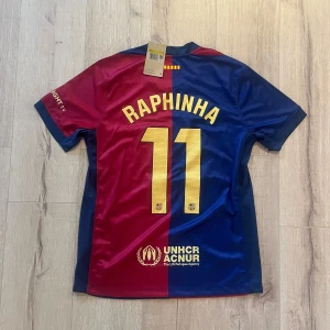 FC Barcelona Raphinha 11 Nike tröja - Säljer en officiell FC Barcelona matchtröja från Nike med Raphinha och nummer 11 på ryggen. Tröjan har klassisk röd och blå färgdelning, guldiga detaljer och klubbmärke på bröstet. Materialet är lätt och ventilerande polyester, perfekt för fotboll eller häng med polarna.