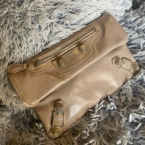 Beige kuvertväska i skinn med detaljer - Snygg beige kuvertväska i skinn med coola spänndetaljer och dragkedja framtill. Väskan har en stilren design och är perfekt att bära i handen. Passar dig som gillar minimalistisk men ändå edgy stil.