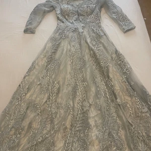 Elegant silvergrå balklänning med spets och paljettdetaljer - Fantastiskt vacker lång balklänning i silvergrå ton, täckt med intrikata spetsbroderier och skimrande paljetter. Klänningen har en figurnära överdel med vadderad byst och markerad midja som övergår i en lätt utsvängd kjol. Detaljer i tyll och spets ger en lyxig, romantisk känsla — perfekt för bal, bröllop, eller en finare fest.