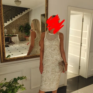 Beige omlottklänning från LIU JO - Supersnygg beige klänning från LIU JO med champagnefärgade broderade detaljer och omlottkänsla. Klänningen är ärmlös, har rund hals och ett unikt mönster som ger en lyxig vibe. Perfekt för dig som vill sticka ut med stilren elegans.