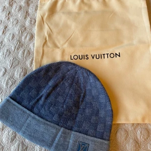 Blå Louis Vuitton mössa i ull - Exklusiv blå mössa från Louis Vuitton med diskret rutig design och vikt kant. Mössan har en broderad LV-logga framtill och är tillverkad i mjuk ullblandning. Perfekt accessoar för kyliga dagar.