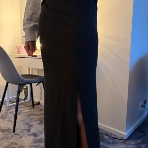 Svart långklänning med slits - Säljer en svart långklänning med stilren design och en snygg slits framtill som ger en cool touch. Klänningen har en rak och figurnära passform som framhäver siluetten. Perfekt för dig som gillar minimalistisk och elegant stil.