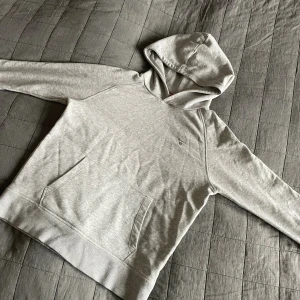 Ljusgrå hoodie från GANT - Snygg ljusgrå hoodie från GANT med broderad logga på bröstet. Klassisk känguruficka framtill, mjuk bomullskänsla och huva. Perfekt för chill dagar eller när du vill ha en clean look.