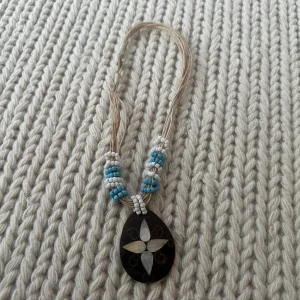 Halsband med pärlor och snäckhänge - Unikt halsband med beige snöre, vita och turkosa pärlor samt ett stort droppformat hänge i mörkt material med inlägg av snäckskal i blommönster. Perfekt för dig som gillar boho-stil och naturmaterial.