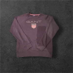 Mörklila sweatshirt från GANT - Säljer en mörklila sweatshirt från GANT med broderad logga och sköld på bröstet. Tröjan har rund halsringning, långa ärmar och ribbade muddar. Perfekt för en chill och stilren look.