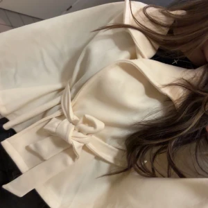 Beige capejacka med bälte och knappar - Snygg beige capejacka med bred krage, knappar framtill och ett matchande bälte i midjan. Jackan har vida ärmar och en stilren, elegant look som passar perfekt till lager-på-lager. Materialet känns mjukt och bekvämt.