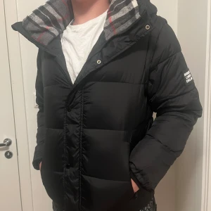 Burberry jacka  - Säljer en svart dunjacka från Burberry London England med avtagbara ärmar så den kan användas som väst. Jackan har rutigt grått innerfoder med röda och svarta detaljer, dragkedja och knappar framtill samt huva. Klassisk Burberry-logga på ärmen. Rikgtigt skön och varm och perfekt inför de kalla tiderna