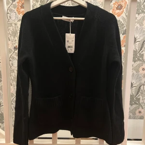 Svart kofta från Wera - Klassisk svart kofta från Wera i mjuk ull. Koftan har två stora fickor framtill, ribbade muddar och stängs med knappar. Perfekt att slänga över en t-shirt eller skjorta för en chill och stilren look, tröjan är i storlek S. Tröjan är enbart testad annars är den som ny🩷