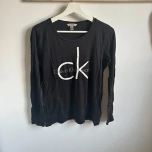 Svart långärmad tröja Calvin Klein - Svart långärmad tröja från Calvin Klein Jeans med stort vitt CK-tryck framtill. Tröjan har rund halsringning och är gjord i mjuk bomull, perfekt för en clean och enkel look. Passar dig som gillar klassiska logoplagg. Trycket är något påverkat, se bild 2.