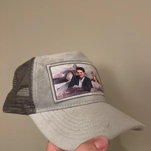 Grå Maggiore keps med mesh och tryck - Säljer en grå keps från Maggiore med mesh på sidorna och baksidan. Framtill finns ett stort tryck med motiv av två personer i en bil och texten 'MAGGIORE'. Kepsen har böjd skärm och justerbar passform. Perfekt för dig som gillar unik streetstyle. Nypris 1000